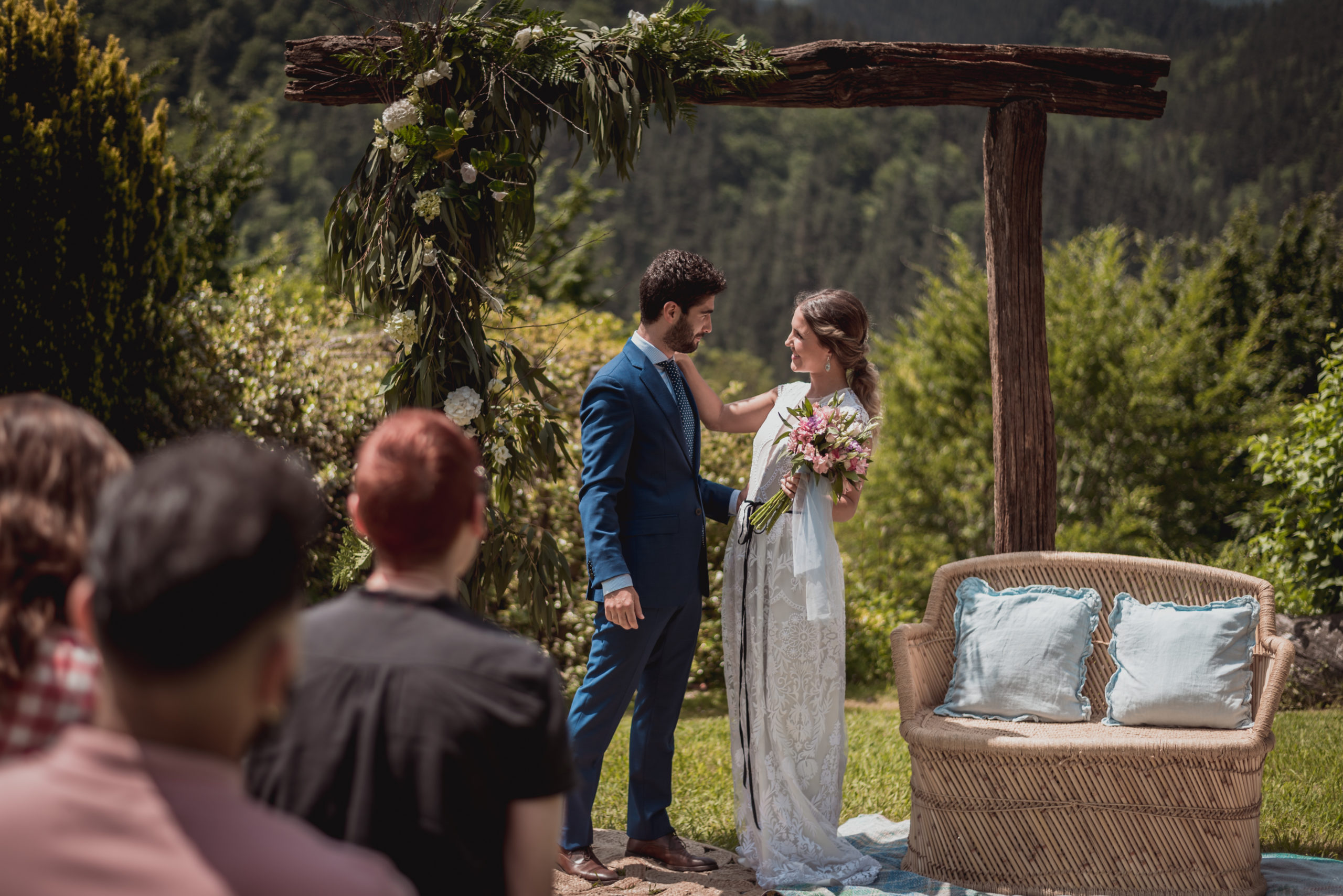 Novia y novio en su ceremonia de boda junto a un sillón doble ratán y un arco natural de fondo