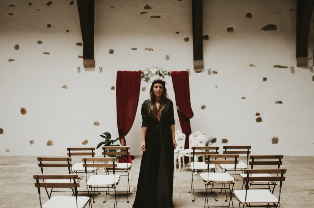 Novia con vestido negro, frente a arco rojo y con sillas de forja y madera al rededor