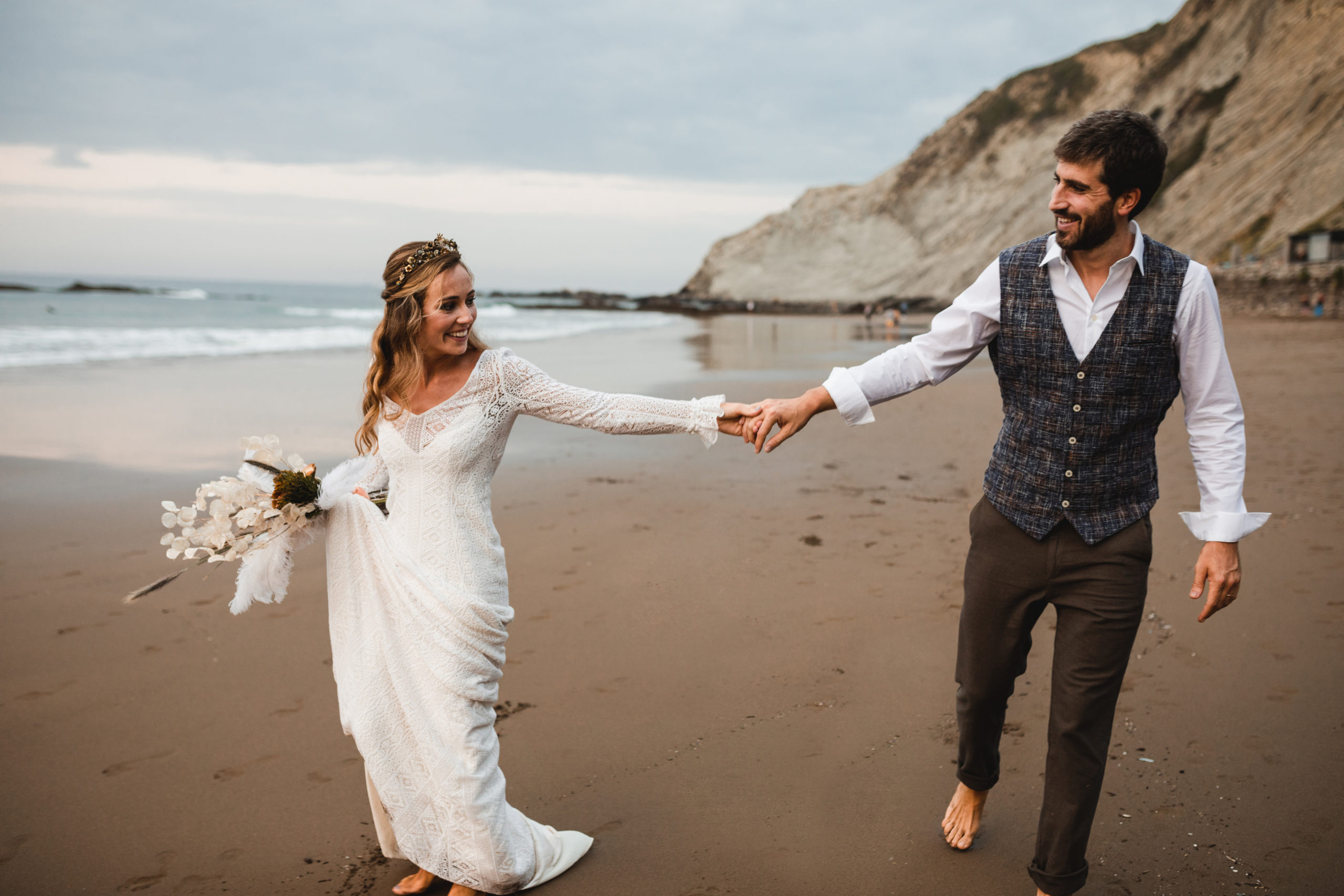 Novio y novia se agarran de la mano paseando por una playa el día de su boda