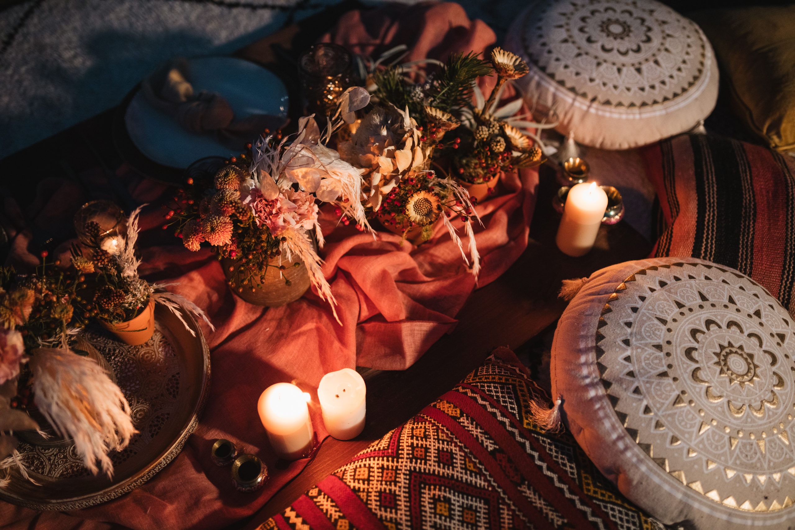 Mesa romántica baja en la playa, con cojines, copas, velas, cubiertos, bajoplatos y flores.