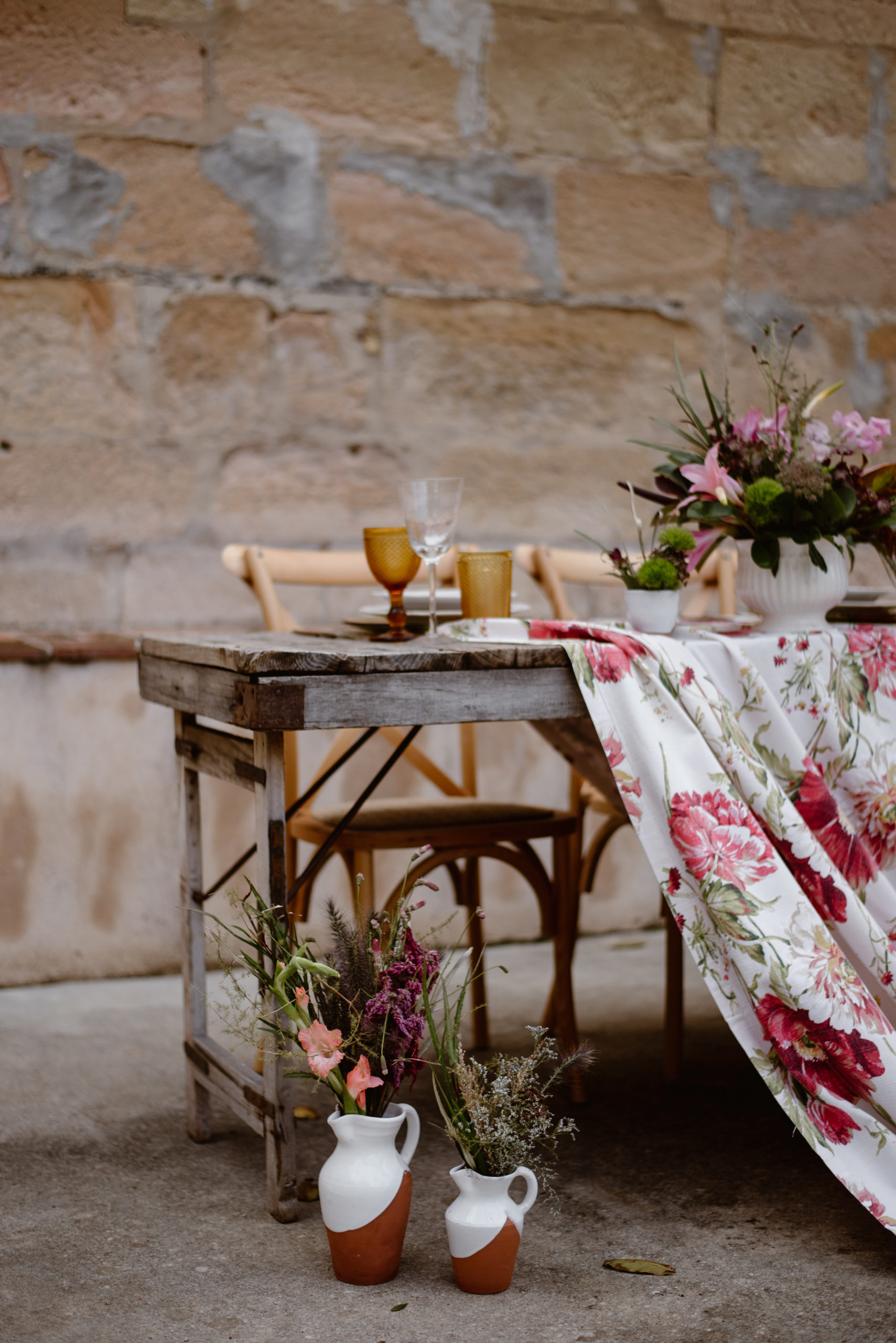 Mesa rústica con mantel floral, vasijas, sillas de madera y copas de colores en alquiler